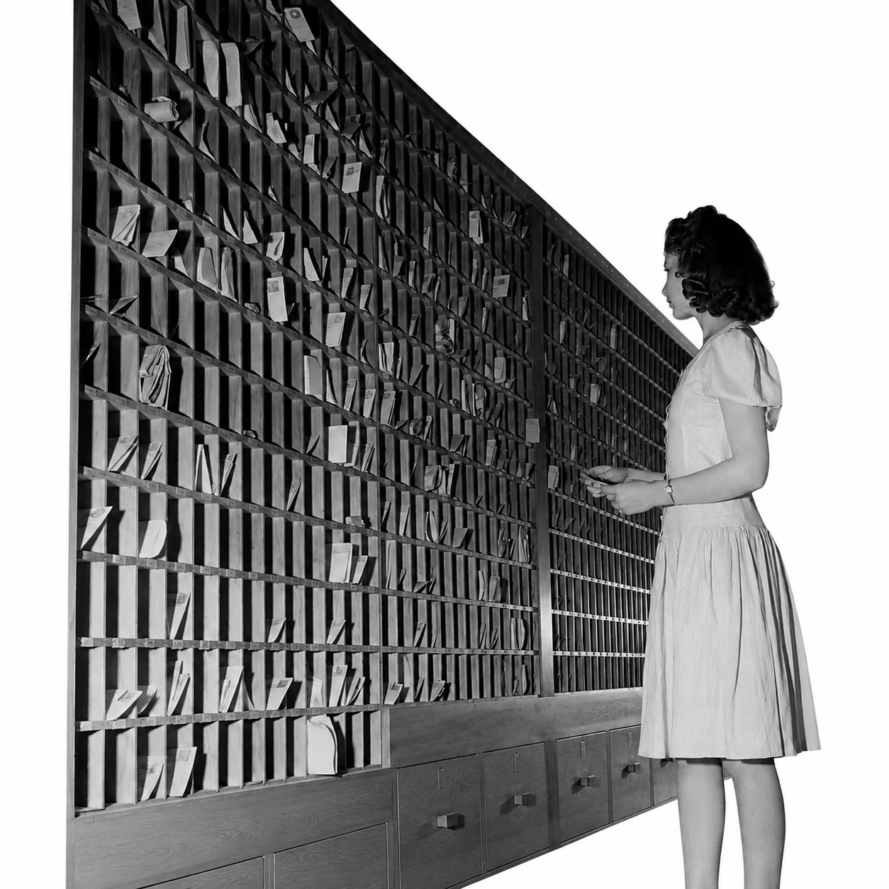 An archive photograph of a clerk at a wall of pigeonhole
card files.