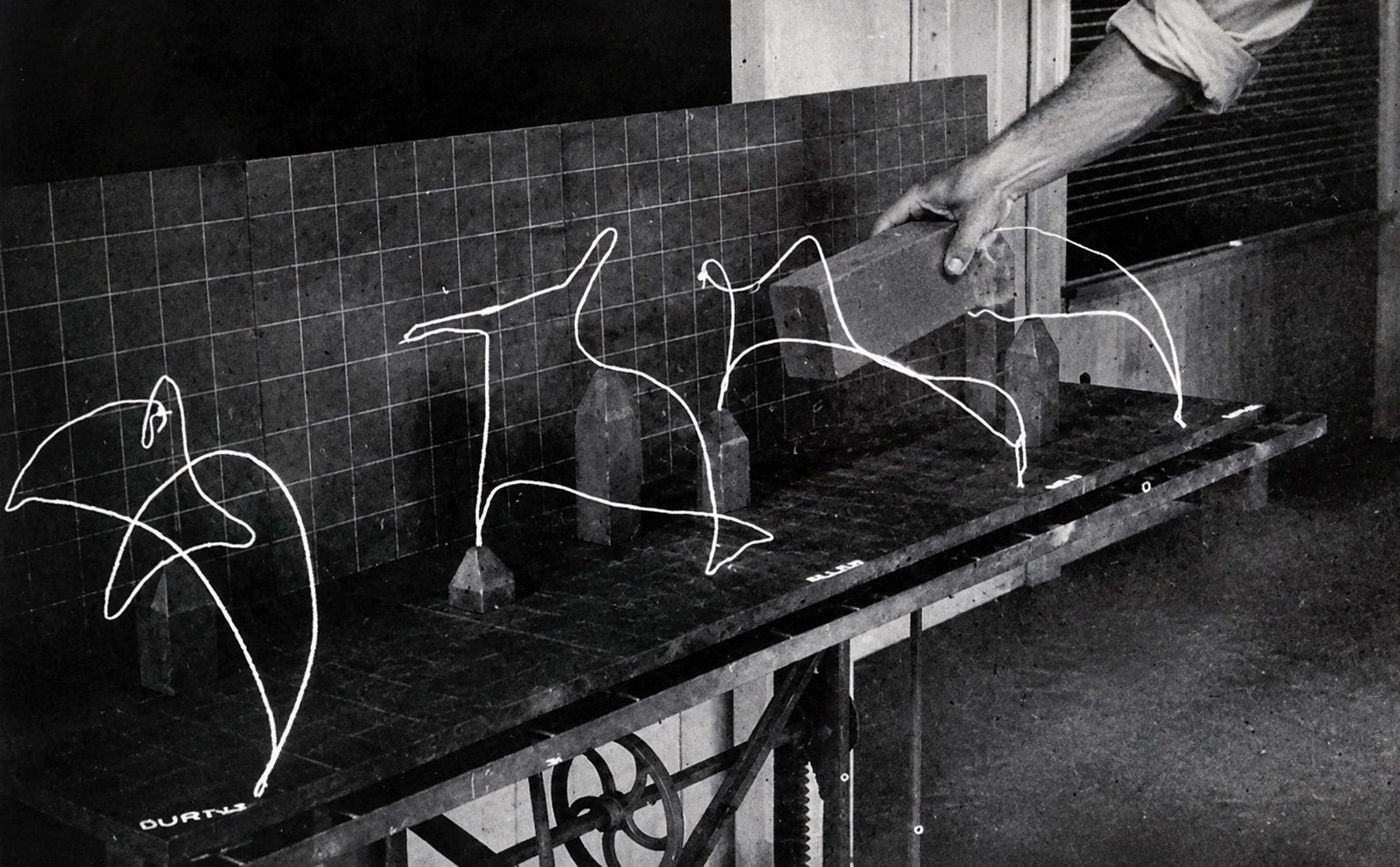 Wire models of a hand's path through the air,
standing in a row on a bench against a gridded backdrop, with a hand lowering a brick
past them.